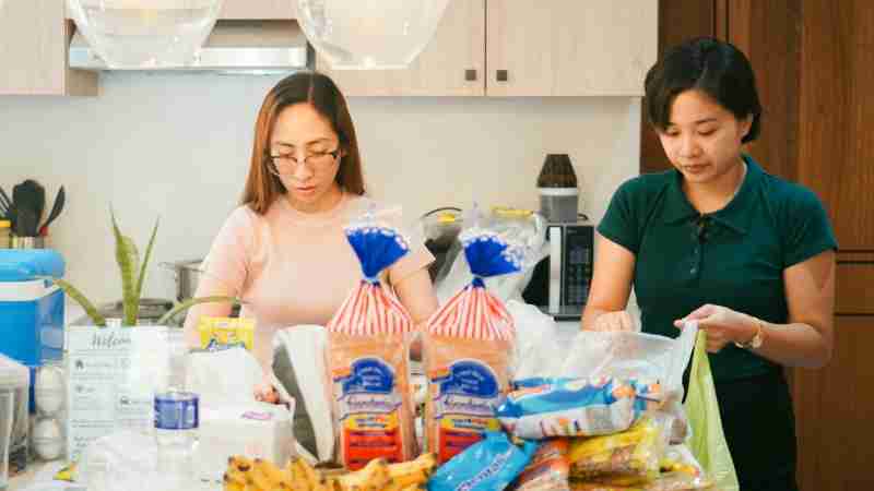 Two women unpack groceries in a modern kitchen setting, featuring bread, bananas, and a variety of packaged foods., tags: climate - pexel